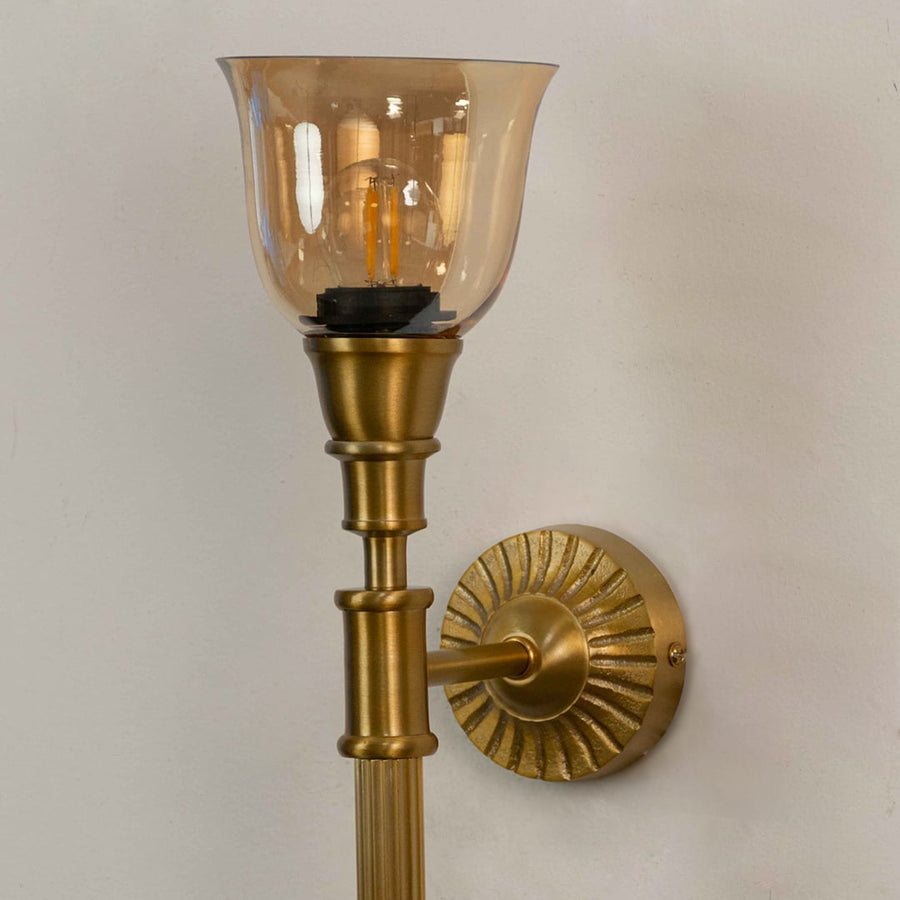 Close up view of brass wall sconce showing ribbed metal arm decorative round backplate and amber glass shade details.