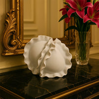 White sculptural ceramic vase featuring dual wave-edged forms placed on a marble tabletop beside a golden ornate mirror.