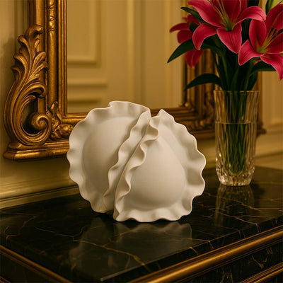 White sculptural ceramic vase featuring dual wave-edged forms placed on a marble tabletop beside a golden ornate mirror.