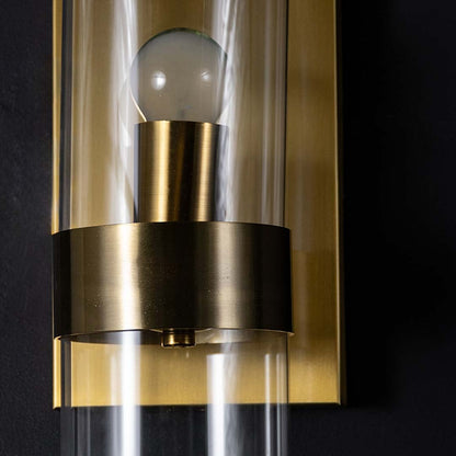 A detailed closeup of the gold wall sconces cylindrical glass shade showing the central bulb and smooth metallic holder with clear reflection and warm tones.
