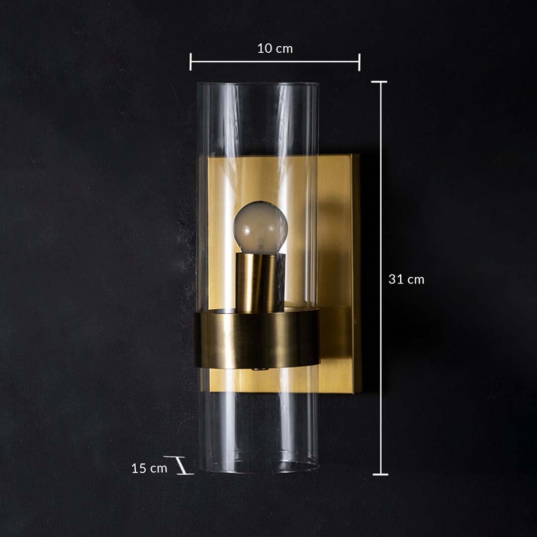 A gold wall light with clear cylindrical glass shade displayed on a black background showing dimensions of 10 cm width 31 cm height and 15 cm depth.