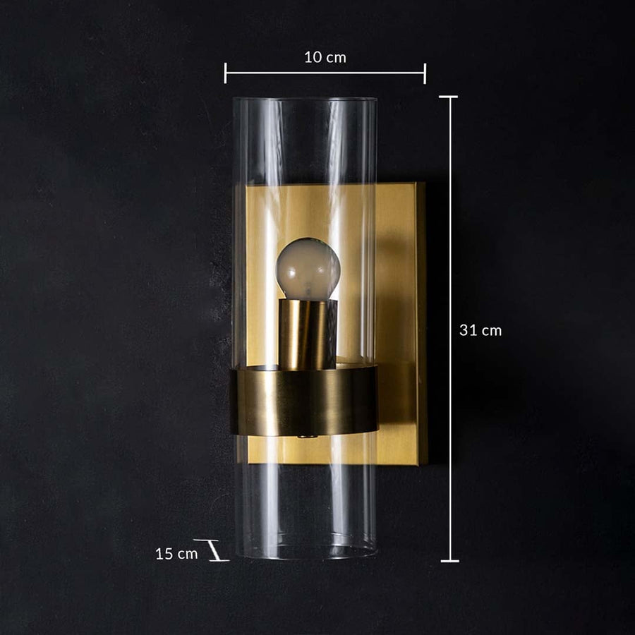A gold wall light with clear cylindrical glass shade displayed on a black background showing dimensions of 10 cm width 31 cm height and 15 cm depth.