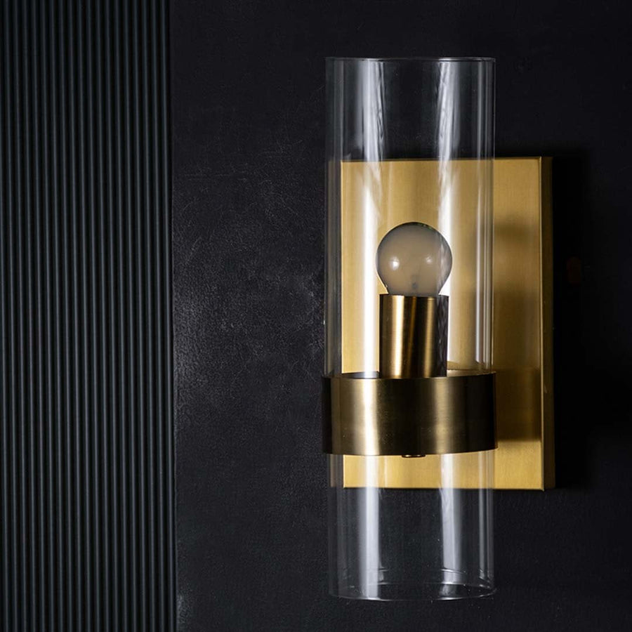 A gold wall light with a tall clear glass shade displayed on a black background with the light off showing the smooth metal details and modern silhouette.