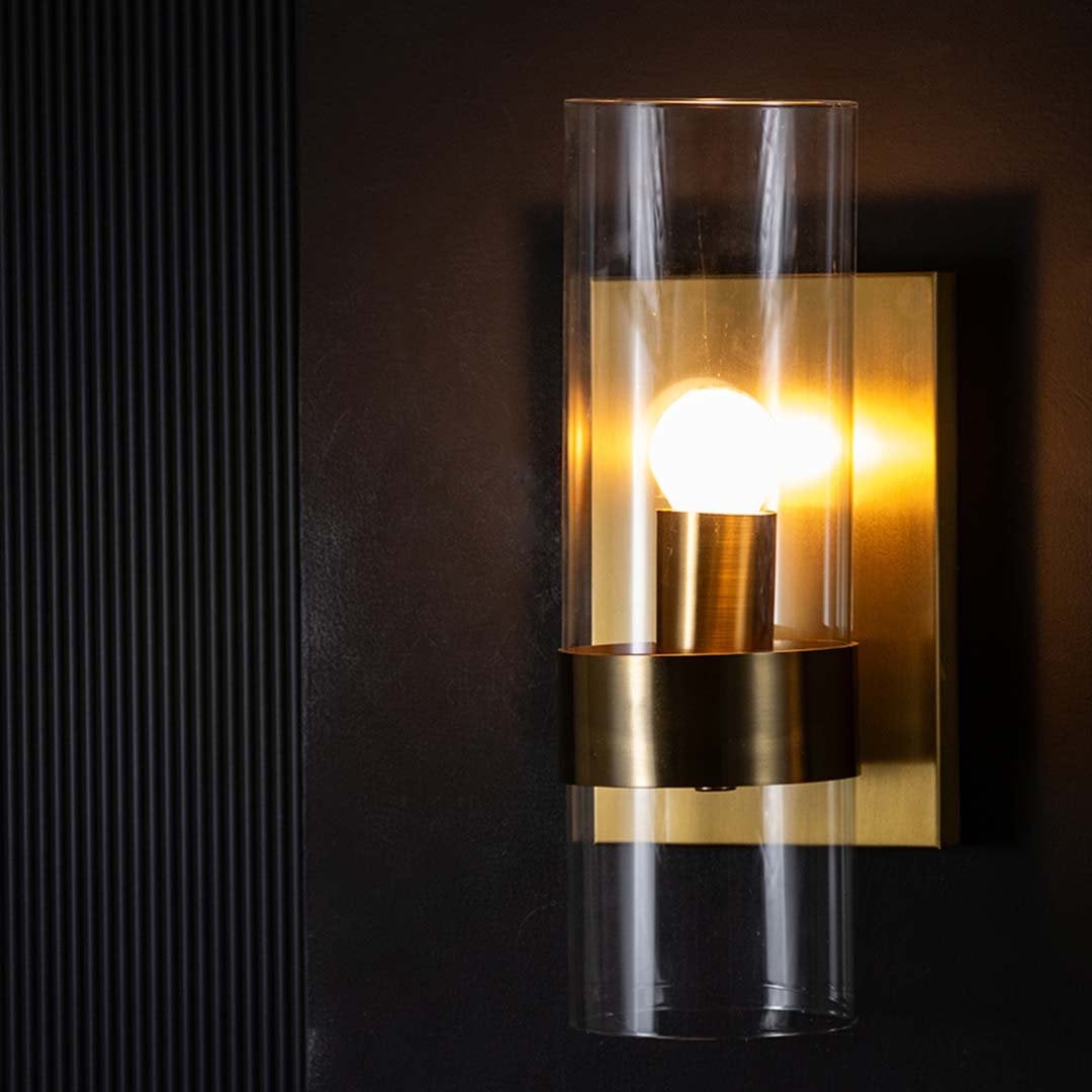 A closeup view of the gold wall light highlighting the transparent glass cylinder and brushed metal base showing refined craftsmanship and modern detailing.