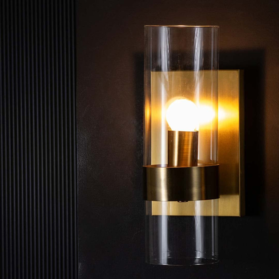 A closeup view of the gold wall light highlighting the transparent glass cylinder and brushed metal base showing refined craftsmanship and modern detailing.