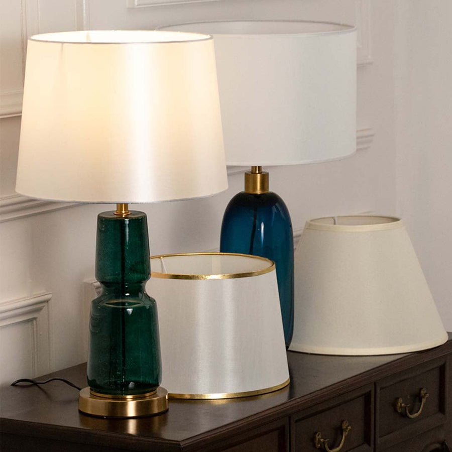White shade with blue and green glass lamps on console table with light on and white wall background for fresh stylish décor.