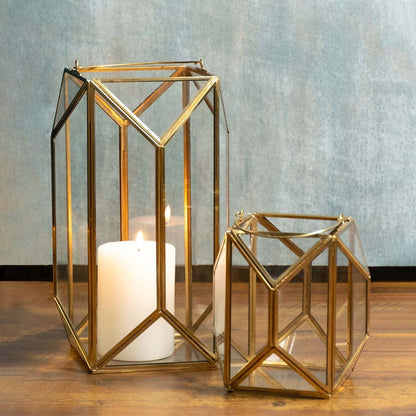 Two gold metal and glass candle lanterns grouped together with candles on creating a warm decorative display perfect for tabletops floors or corners.