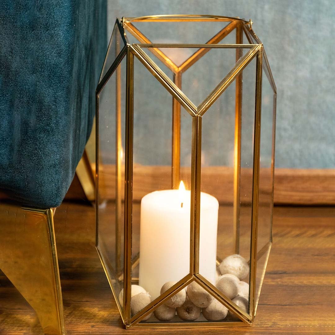 Elegant tall gold metal and clear glass candle lantern placed beside a teal chair with the candle lit adding a warm decorative touch to modern home interiors.