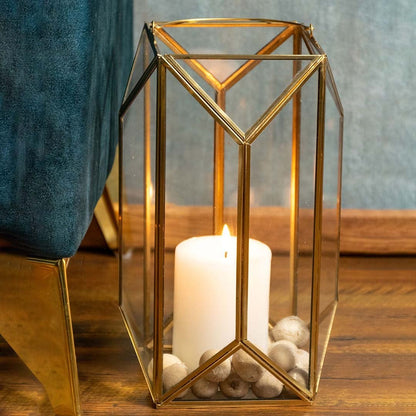 Elegant tall gold metal and clear glass candle lantern placed beside a teal chair with the candle lit adding a warm decorative touch to modern home interiors.