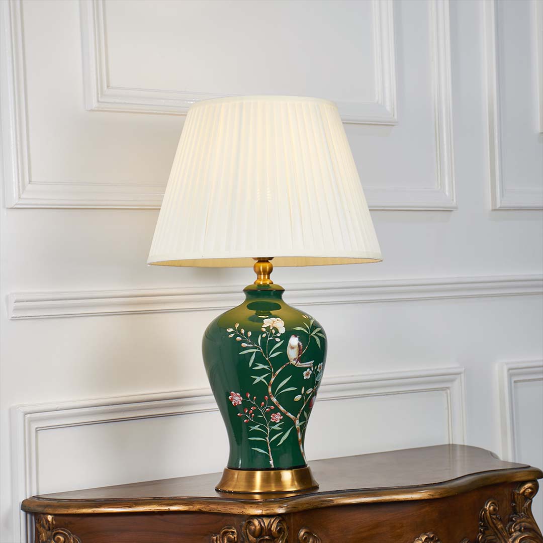 Green ceramic floral table lamp shown with light on against a white background highlighting its warm illumination and artistic hand painted details.