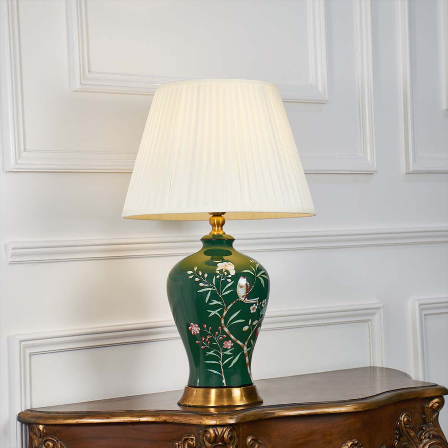 Green ceramic floral table lamp shown with light on against a white background highlighting its warm illumination and artistic hand painted details.