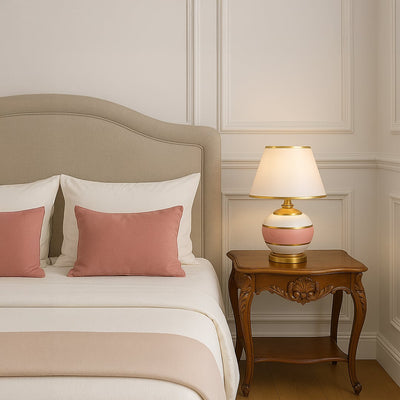 Pink ceramic table lamp with gold accents and white shade glowing on bedside table for elegant bedroom lighting decor.