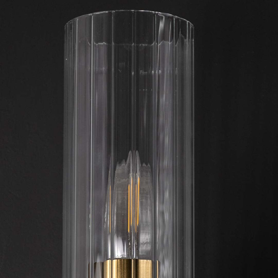 A detailed closeup view of the gold wall light showing the ribbed clear glass shade and polished metal arm with the bulb glowing softly creating a warm ambient effect.
