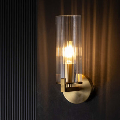 A glowing ribbed glass shade wall sconce with a gold base displayed on a black background with the light on for a dramatic warm effect.