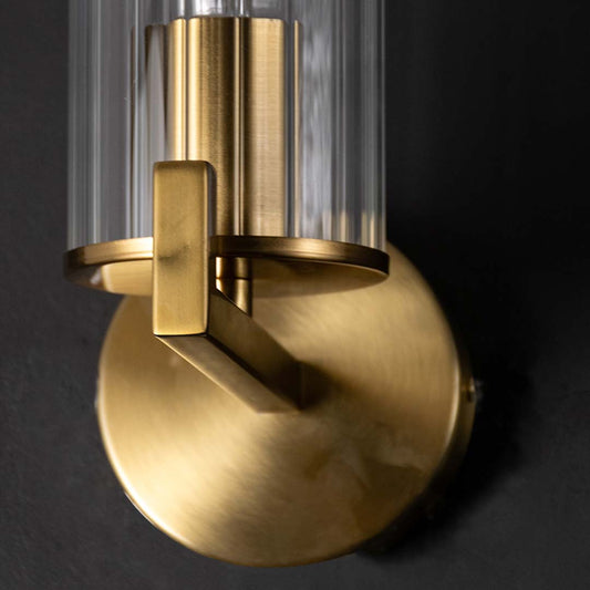 Close up details of a premium gold brass wall sconce lamp base with clear glass rod shade over dark textured backdrop.