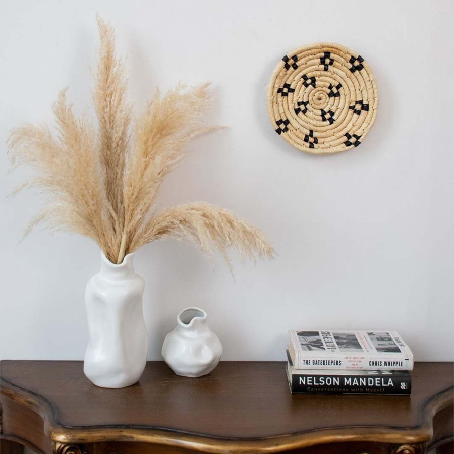 Woven wall basket styled on a white wall above console table with pampas grass and books.