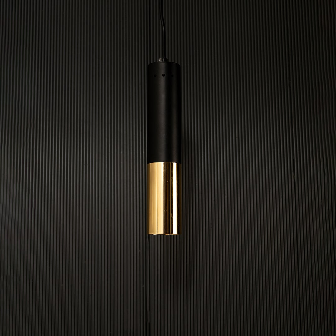 Black and gold cylinder pendant light hanging against dark textured wall with light on for minimalist modern décor.