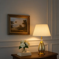 Aurora glass table lamp with pleated shade and golden base styled in living room with light turned on for warm ambience.