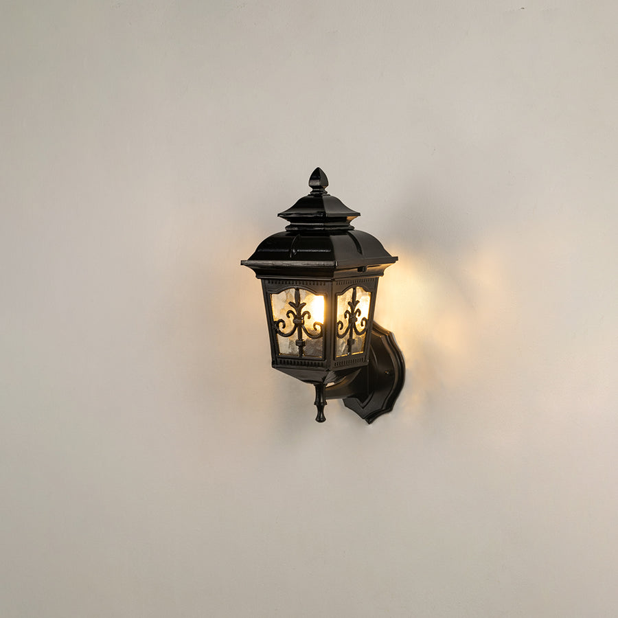 Classic black outdoor wall lantern on white background with clear glass panels and intricate metal overlay filigree decor.