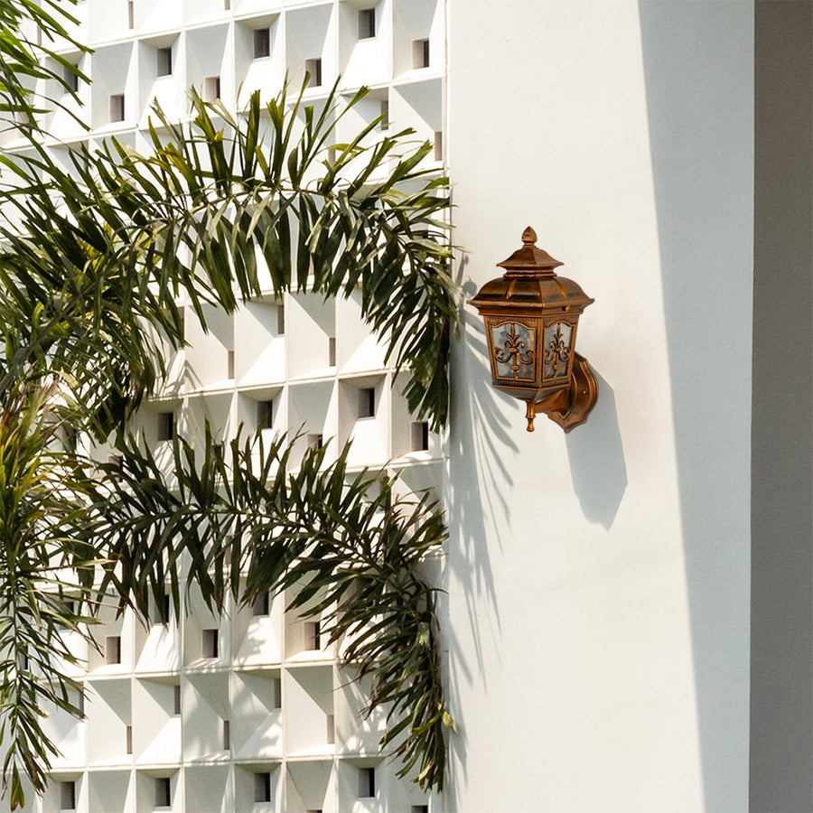 A gold outdoor wall lantern mounted on a white exterior facade adding decorative charm and subtle illumination to a bright open outdoor space in natural daylight.