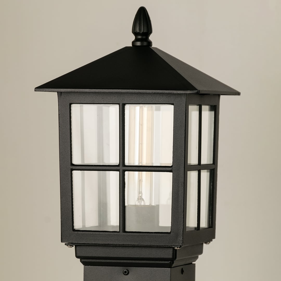 Closeup of black outdoor pillar lamp showing clear framed glass and warm filament bulb detail suitable for elegant garden and pathway lighting.