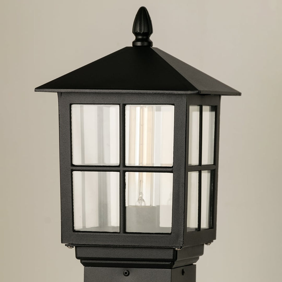 Closeup of black outdoor pillar lamp showing clear framed glass and warm filament bulb detail suitable for elegant garden and pathway lighting.
