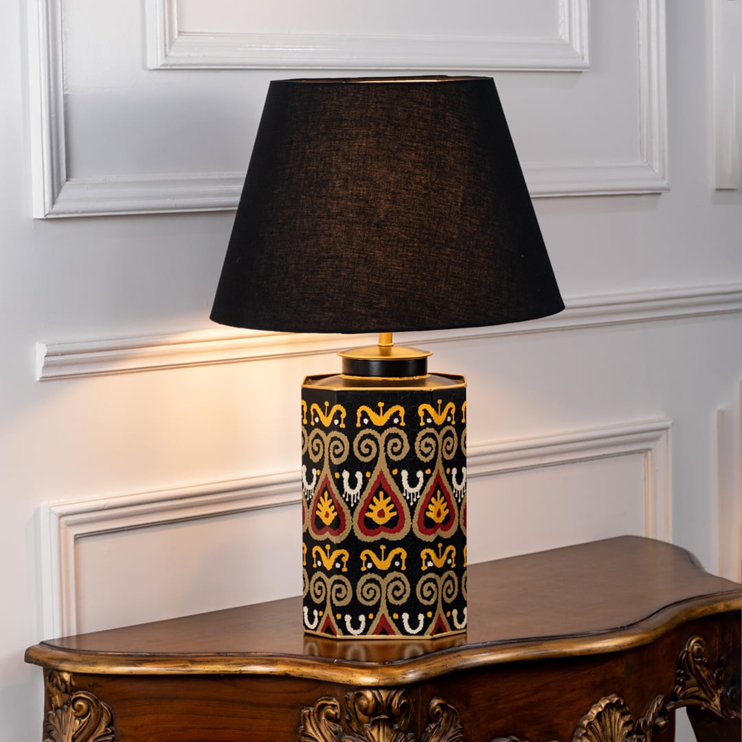 Table lamp on white background featuring black and gold geometric patterned ceramic base with dark fabric shade and warm ambient glow.