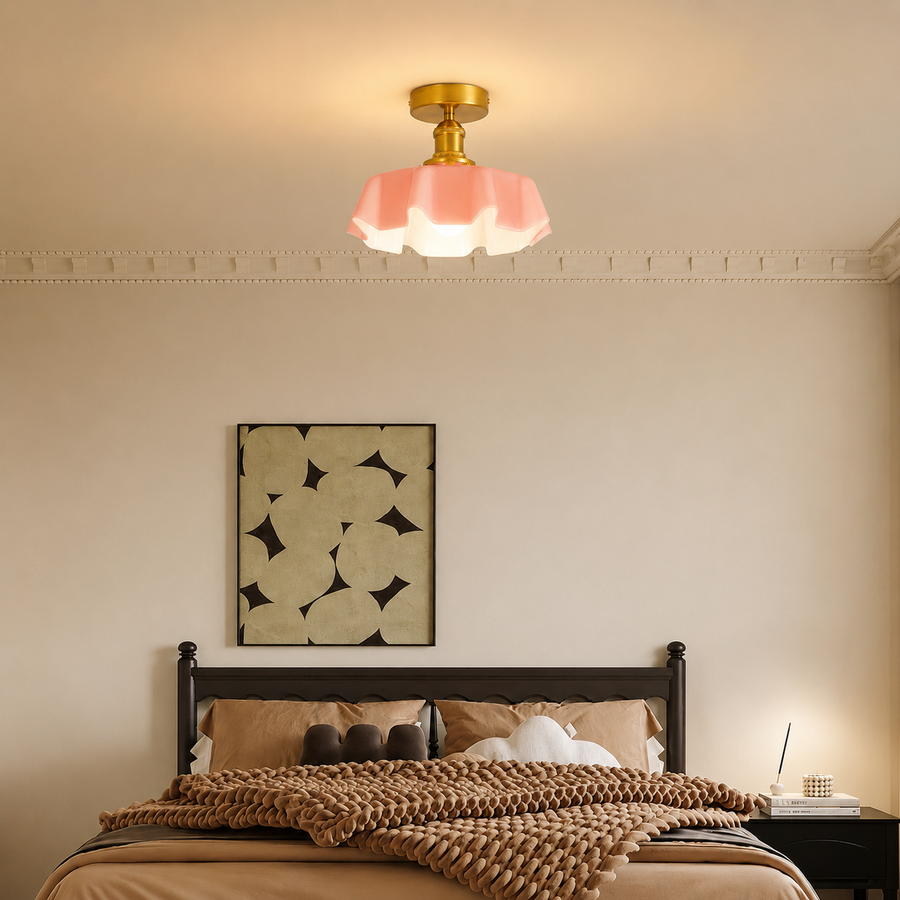 A modern ceiling light fixture with a pink flower-shaped shade and a gold brass ceiling mount, shown illuminated in a warm bedroom setting above a bed.