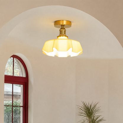 A semi-flush ceiling light fixture with a warm yellow glass shade and a gold-toned brass ceiling mount, shown installed in an arched interior space with a red-framed window and a plant.