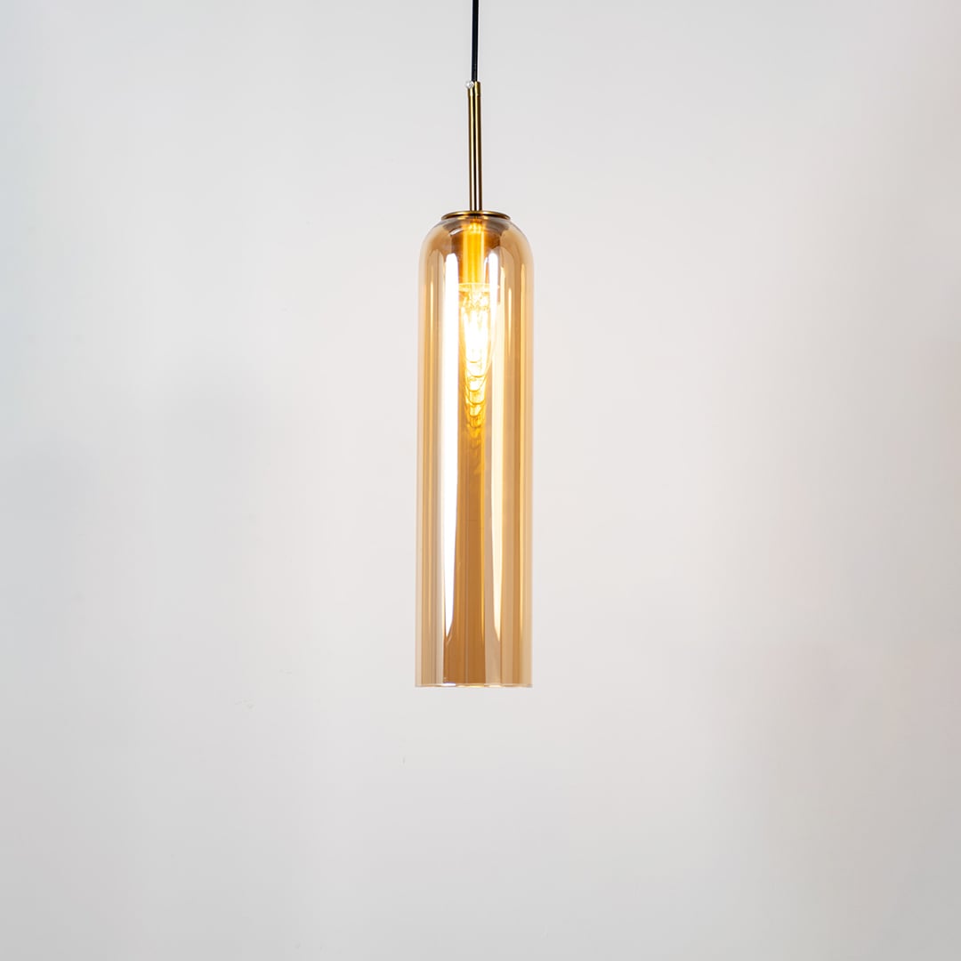 A tall cylindrical pendant light with its bulb glowing warmly on a white background showing its smooth transparent glass body and elegant metal detailing with the light on.