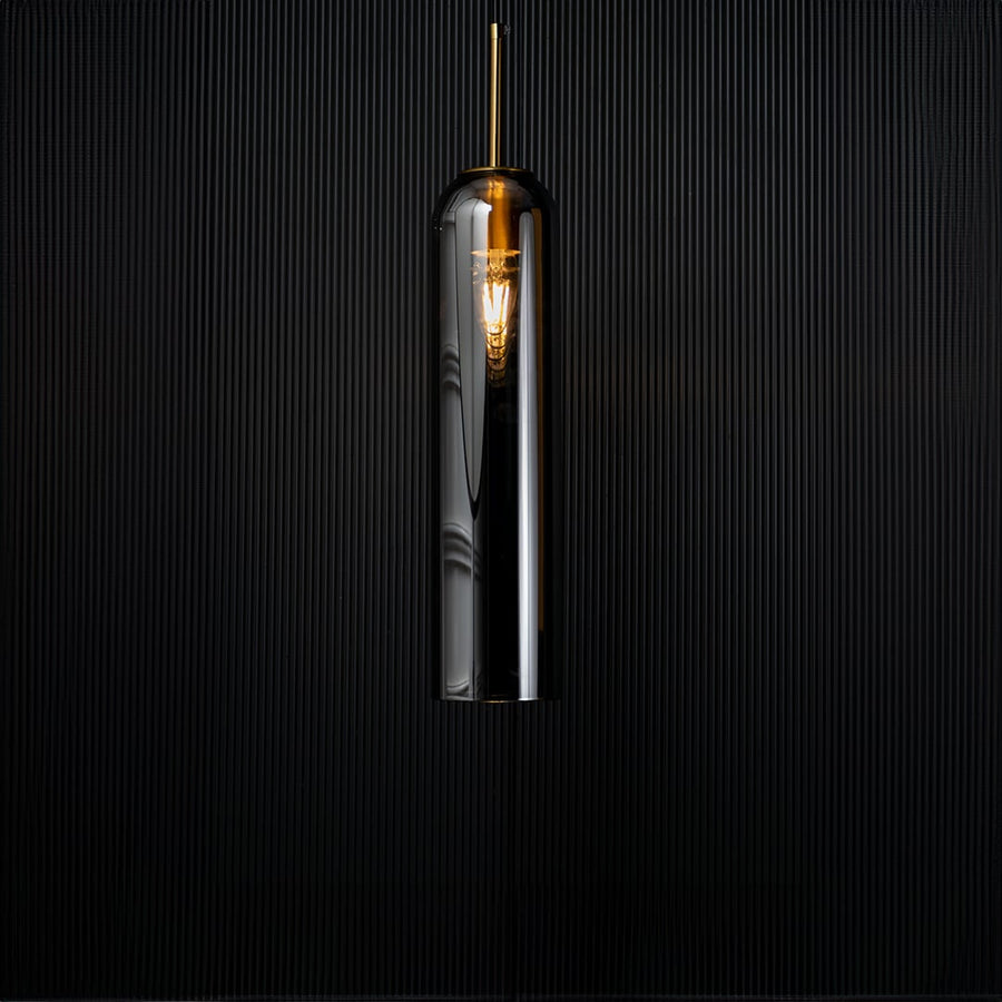 A modern silver cylindrical pendant light against a deep black background with the light turned on casting a warm glow and highlighting its reflective finish.
