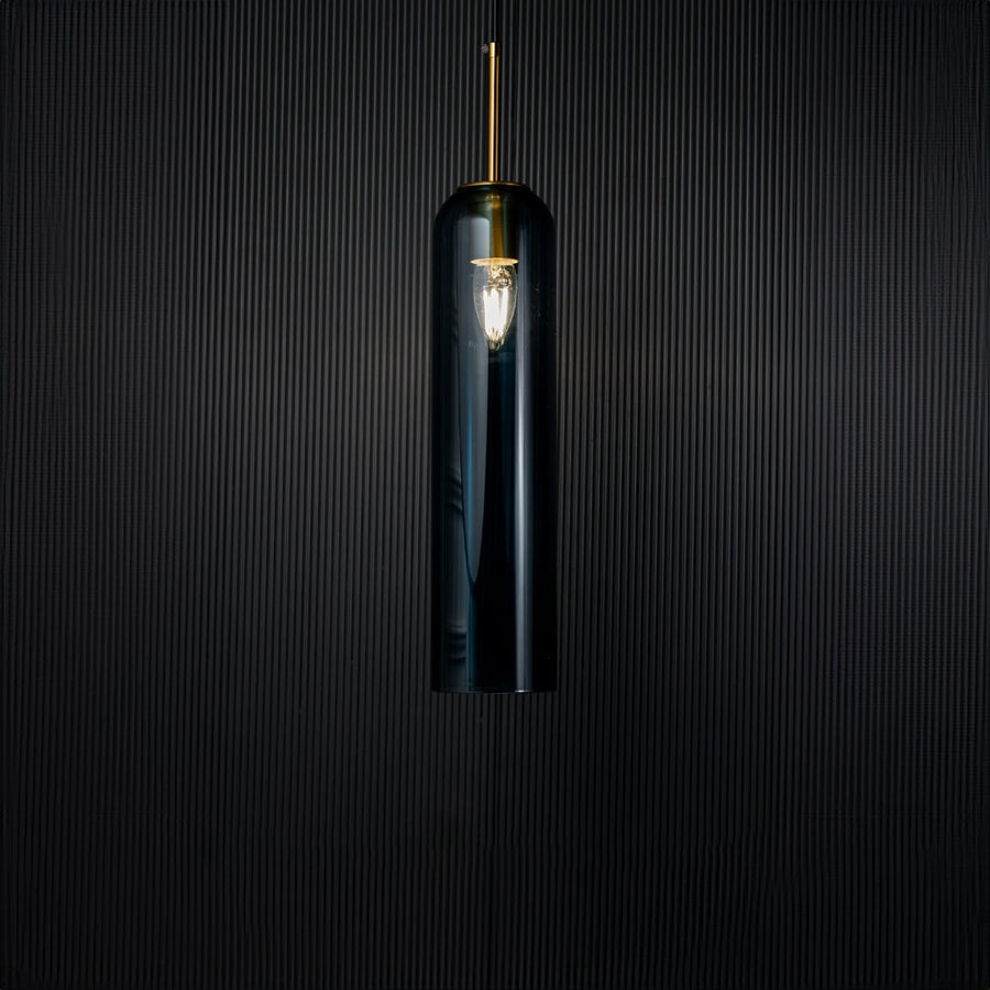 A glowing blue glass pendant light turned on against a deep black background creating a striking contrast that enhances its tall transparent design and warm radiance.