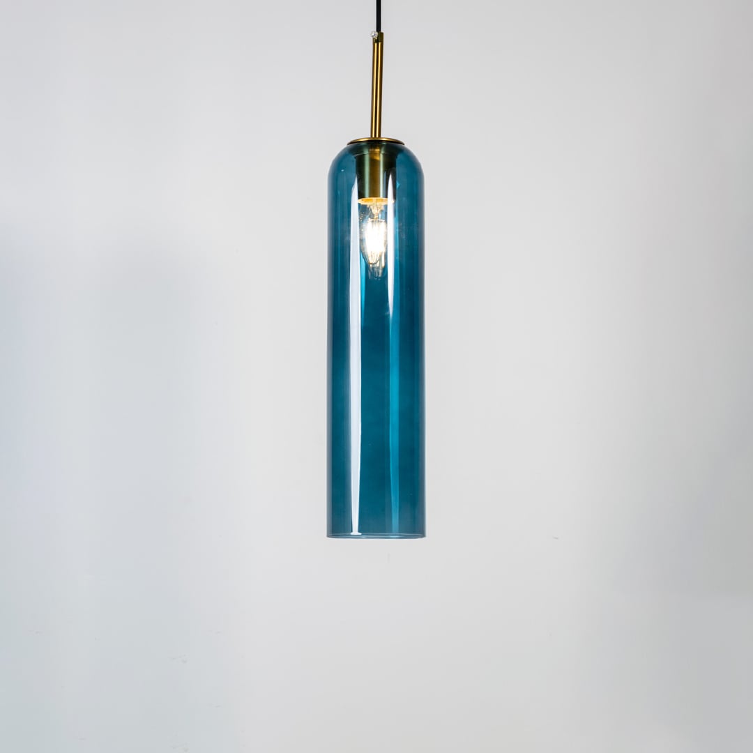 A blue glass pendant light turned on against a clean white background showcasing its glowing warm illumination and tall cylindrical design with gold detailing.