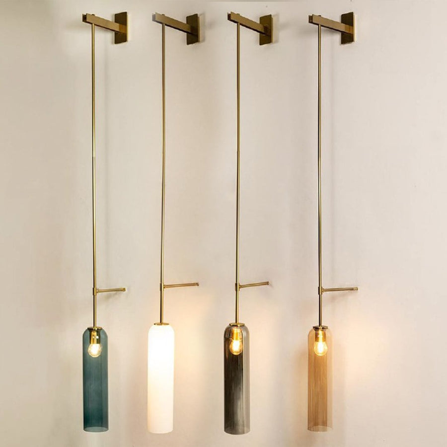 Four pendant lights with long gold rods and tall cylindrical shades in blue white smoky grey and amber hanging in a row and glowing softly against a light wall.