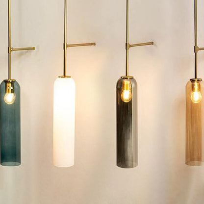 Close up of four cylindrical pendant lights with long glass shades in blue white grey and amber hanging from slim gold rods each with a glowing bulb.
