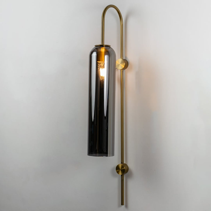 A tall glass wall light with metal frame shown with the light on against a clean white background highlighting its warm glow and modern structure.