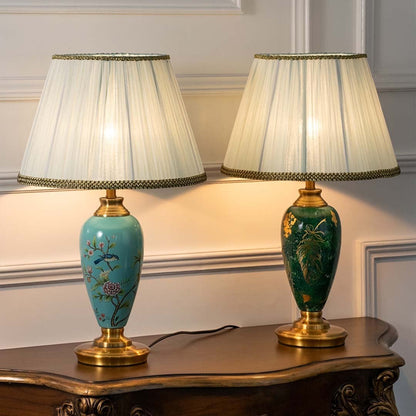 Turquoise ceramic table lamp with floral hand-painted design and pleated white shade turned on against white background creating warm soft lighting.