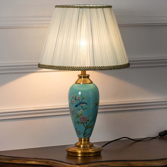 Turquoise ceramic table lamp with floral hand-painted design and pleated white shade turned on against white background creating warm soft lighting.