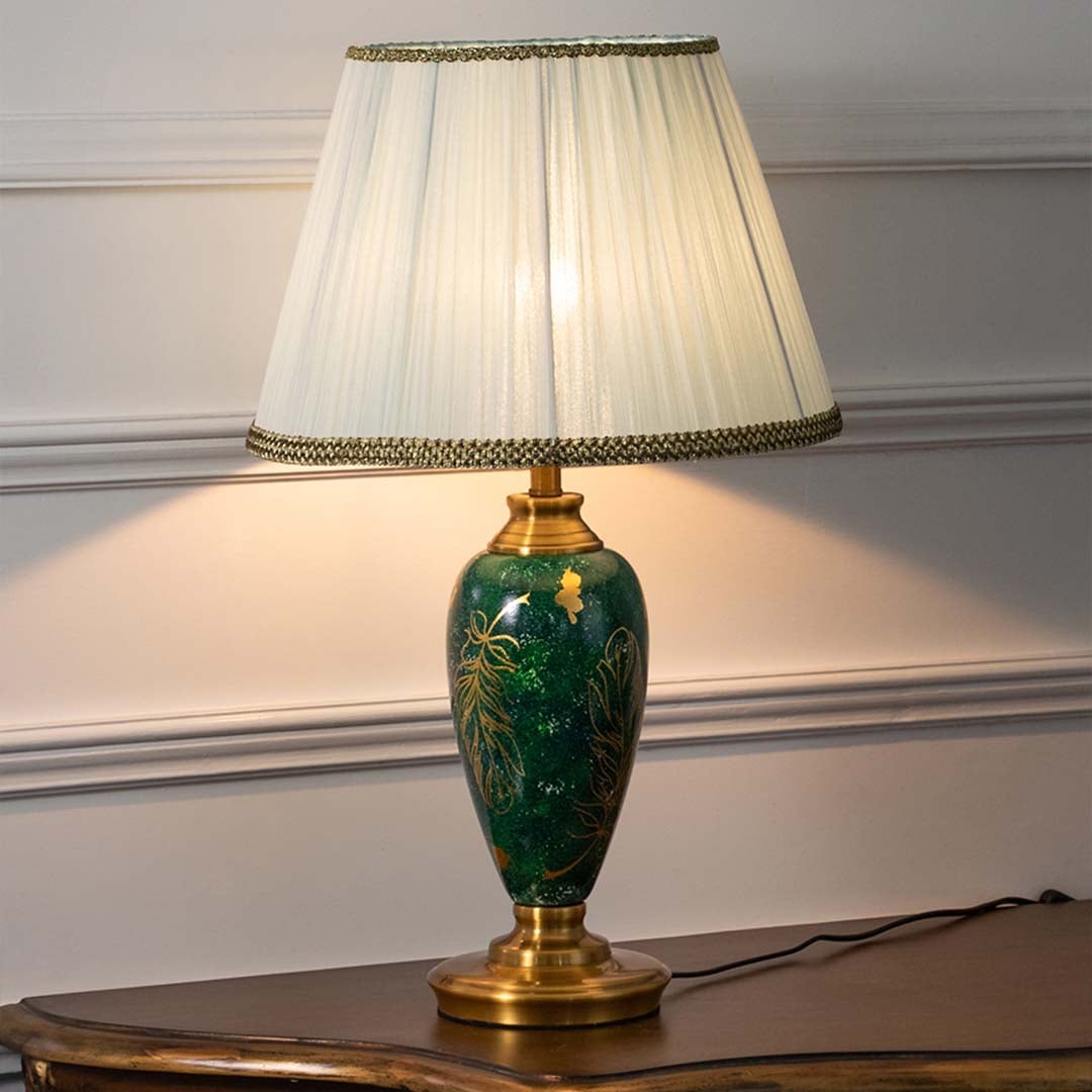 Emerald green ceramic table lamp with golden leaf motifs and pleated white shade glowing warmly on a white background creating a refined and luxurious appeal.
