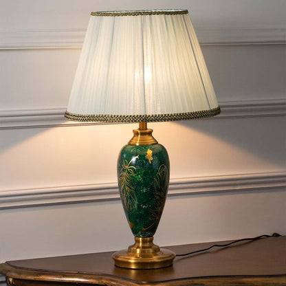 Emerald green ceramic table lamp with golden leaf motifs and pleated white shade glowing warmly on a white background creating a refined and luxurious appeal.