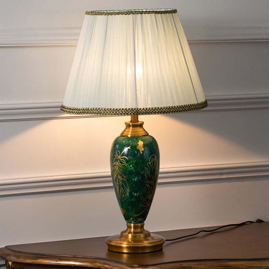 Emerald green ceramic table lamp with golden leaf motifs and pleated white shade glowing warmly on a white background creating a refined and luxurious appeal.