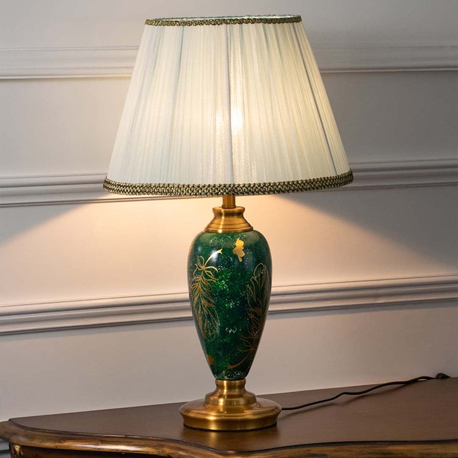 Emerald green ceramic table lamp with golden leaf motifs and pleated white shade glowing warmly on a white background creating a refined and luxurious appeal.