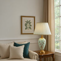 Elegant mint green ceramic table lamp with golden base and hand-painted floral motifs glowing warmly beside beige sofa creating cozy sophisticated living room ambiance.