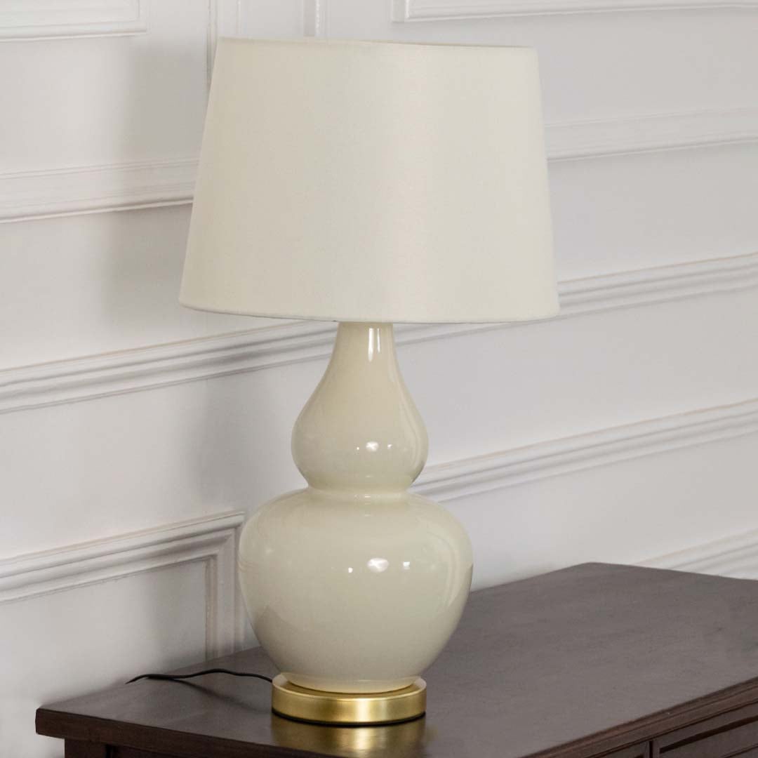 White shade beige ceramic table lamp on wooden side table with light off and white panel wall for clean modern décor.