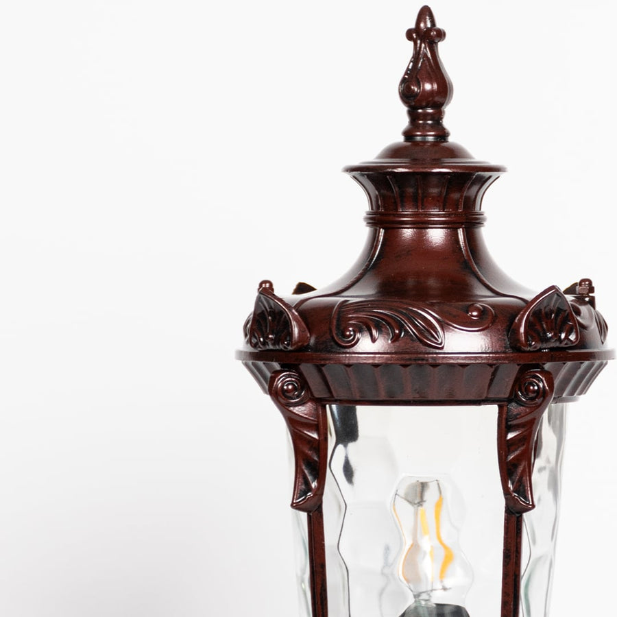Closeup of antique brown pillar light showing carved frame detailing and patterned clear glass for elegant outdoor lighting.