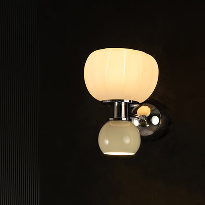 Modern wall sconce turned on displaying pumpkin shape white glass and round beige glass shades with chrome metal body on plain black background.