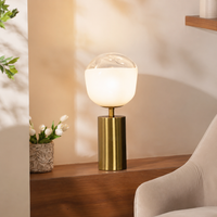 Luxury gold brass cylinder table lamp with a glowing frosted white round glass globe setup on a wooden side cabinet.