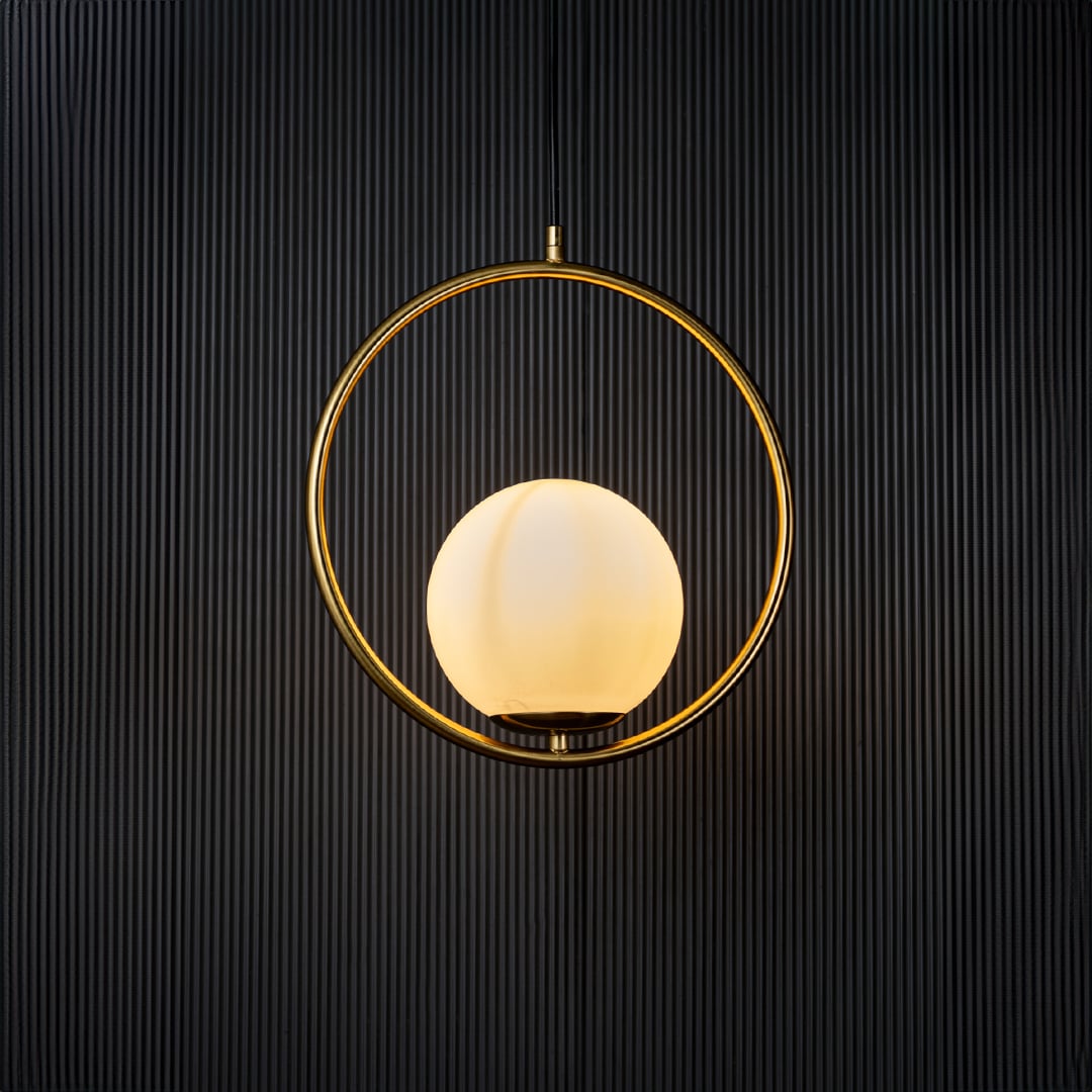 A circular pendant lamp illuminated against a dark black background creating a warm ambient glow that highlights the golden ring and smooth white globe.