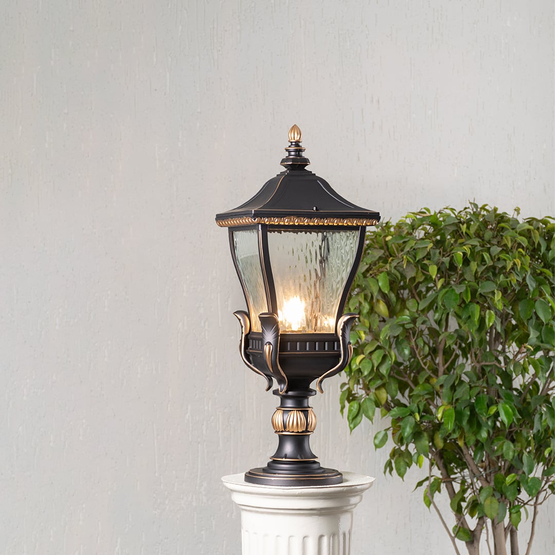 Elegant black and gold metal outdoor pillar light glowing with clear glass shade placed on decorative column lifestyle view.