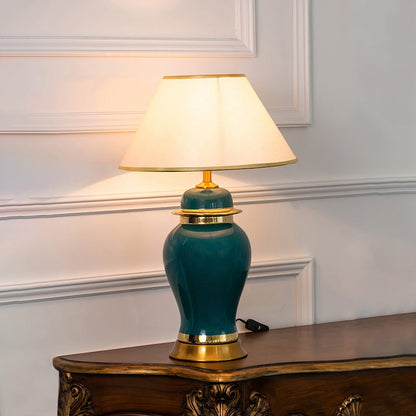 Green ceramic lamp with golden base and white shade turned on glowing against a plain white background.